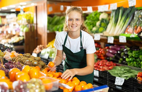 A smiling grocery worker in a green apron arranges oranges in a produce display.