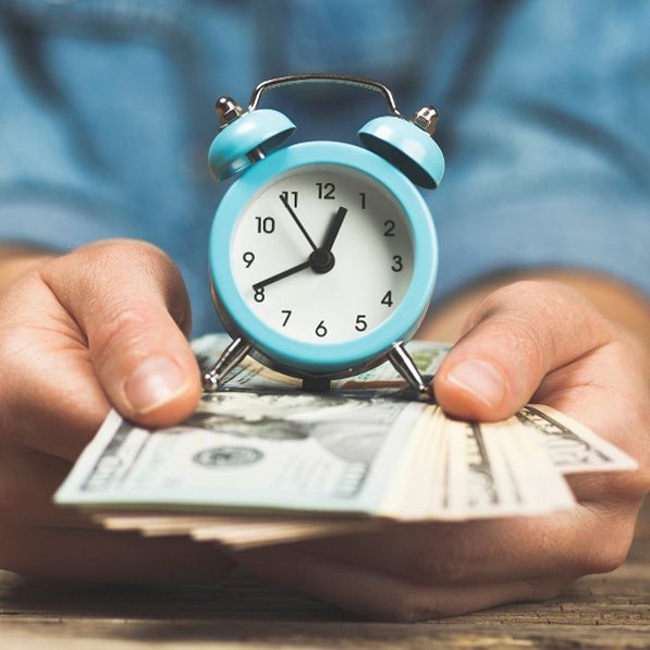 Hands hold U.S. banknotes with a small blue alarm clock resting on top.