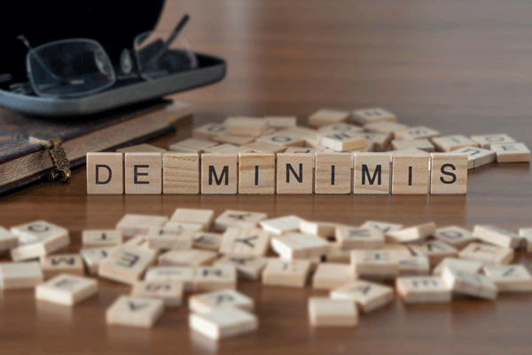 Wooden letter tiles spell DE MINIMIS on a desk among scattered tiles and glasses.