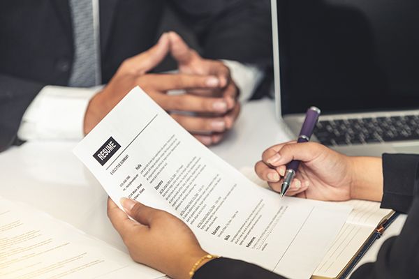 An interviewer holds a résumé and pen across a desk from a seated applicant.