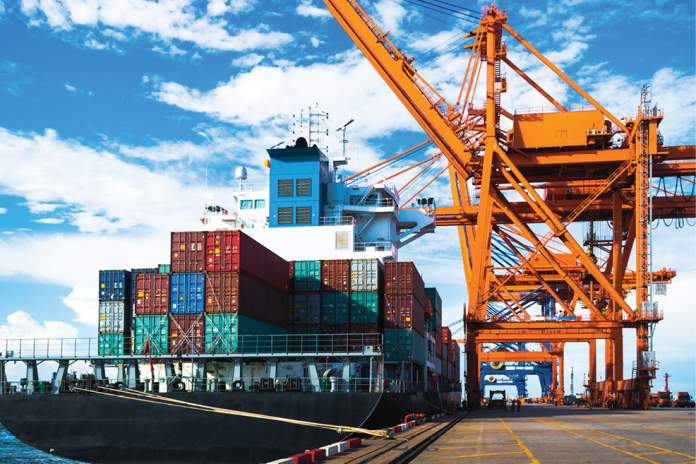 A container ship sits beside large orange port cranes beneath a blue sky.