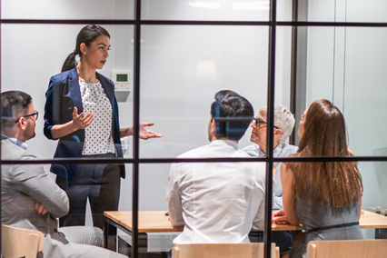 A woman leads a meeting with seated coworkers behind a glass office wall.