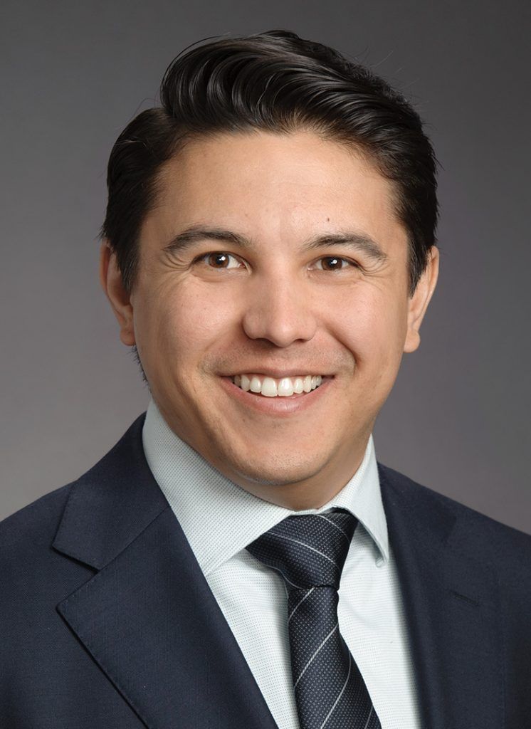 Portrait of a smiling man in a dark suit, white shirt and striped tie against gray.