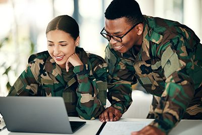Two people in military camouflage smile while reviewing a laptop together.