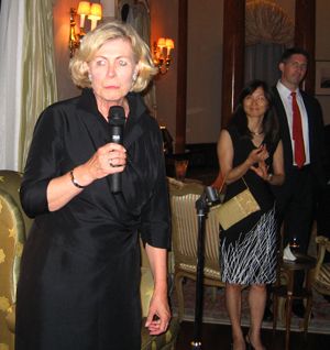 A woman in black speaks into a handheld microphone at an indoor gathering.