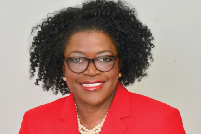 Portrait of a smiling woman with curly hair and glasses wearing a red blazer and pearl necklace.