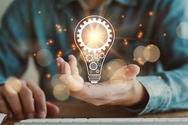 A glowing lightbulb with gears appears above a person's open hand beside a laptop.