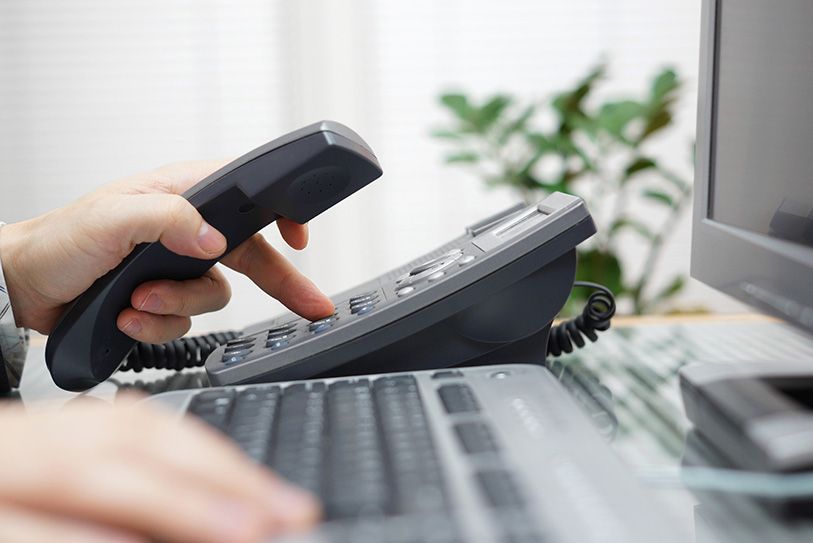 A hand lifts a desk telephone receiver beside a laptop.