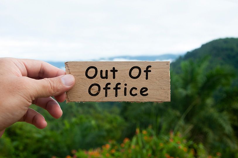 A hand holds a cardboard sign reading Out Of Office against a green mountain landscape.