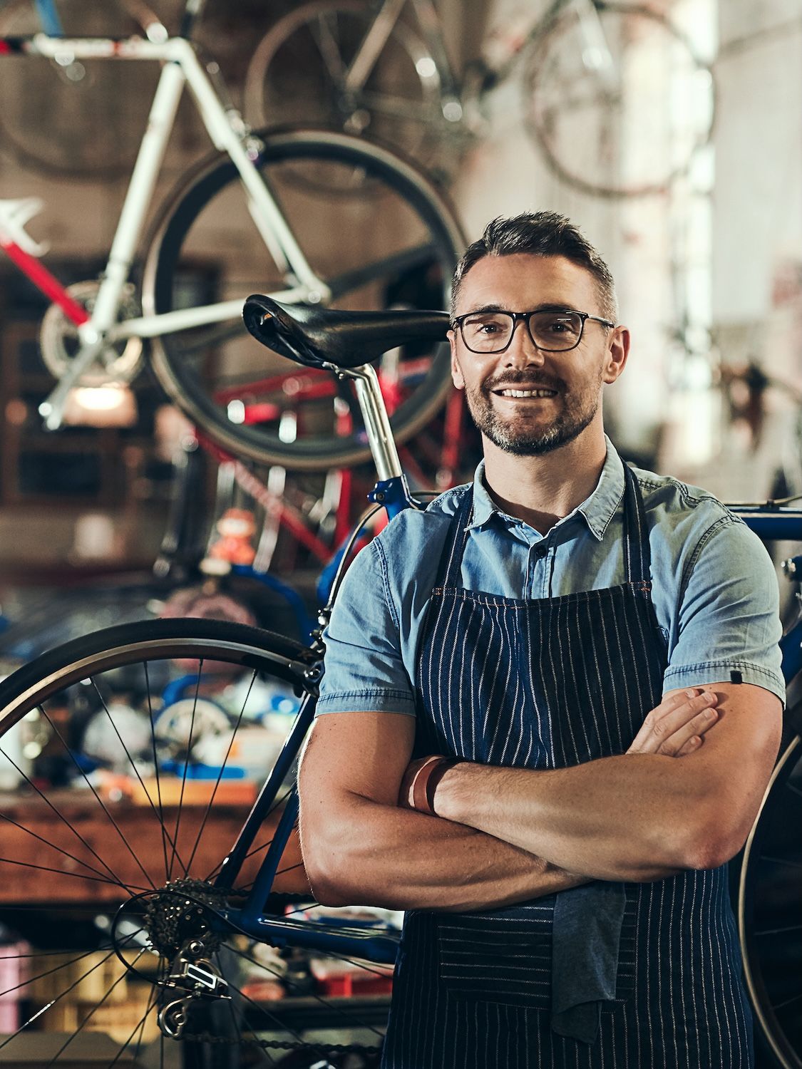 A smiling bicycle shop worker in an apron stands with folded arms among bicycles.