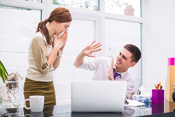 A woman covers her mouth while a seated coworker raises his hands beside a laptop.