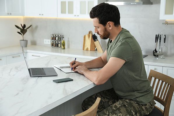 Man in a green shirt and camouflage pants writing in a notebook at a kitchen counter, with a laptop and phone nearby.