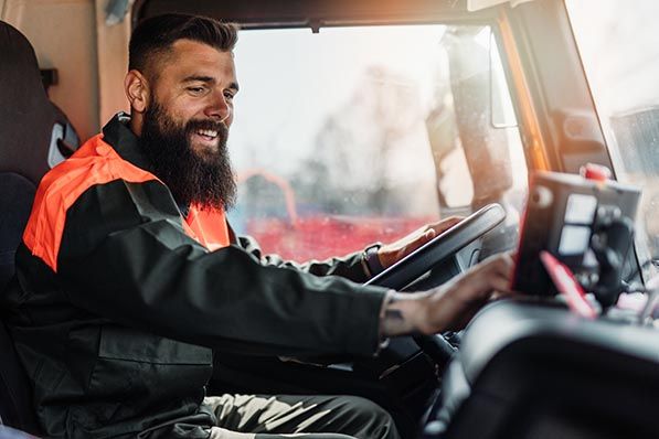 A smiling bearded driver in work clothes sits behind the wheel of a truck.