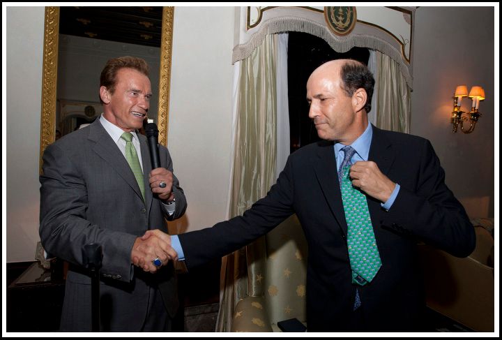 Two men in suits shake hands in an ornate room, one holding a microphone.