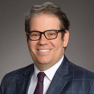 Portrait of a smiling man with rectangular glasses, a checked navy suit and burgundy tie.