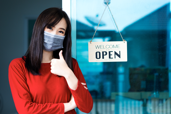 A masked shop worker gives a thumbs-up beside a Welcome Open sign.