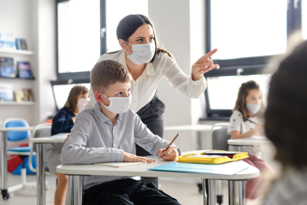 A masked teacher points across a classroom while helping a masked student at a desk.
