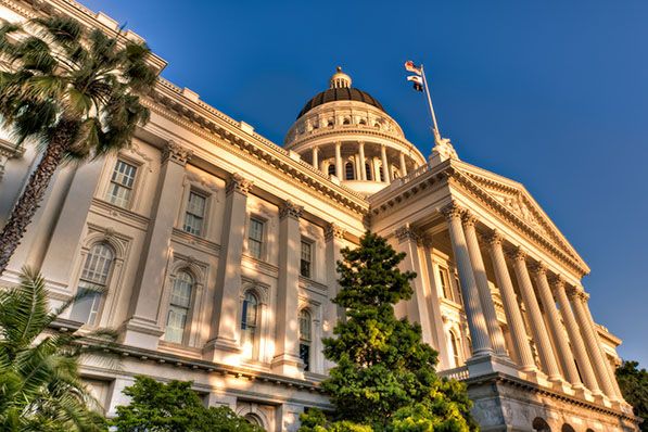 Low-angle view of the California State Capitol facade and dome beside palm trees.
