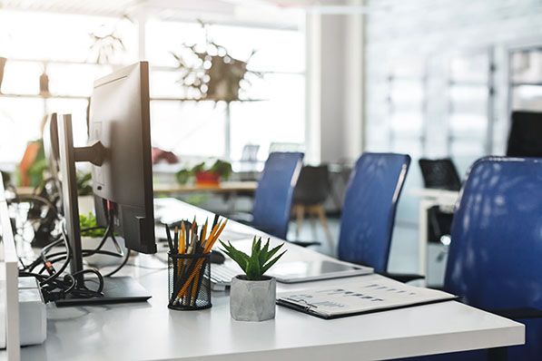 An empty office workstation with a monitor, desk supplies, small plant and blue chairs.