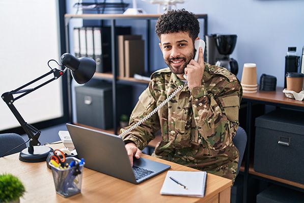 A smiling man in military camouflage uses a phone beside a laptop and desktop microphone.