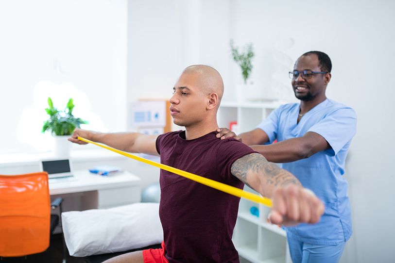 A physical therapist assists a seated person stretching a yellow resistance band.