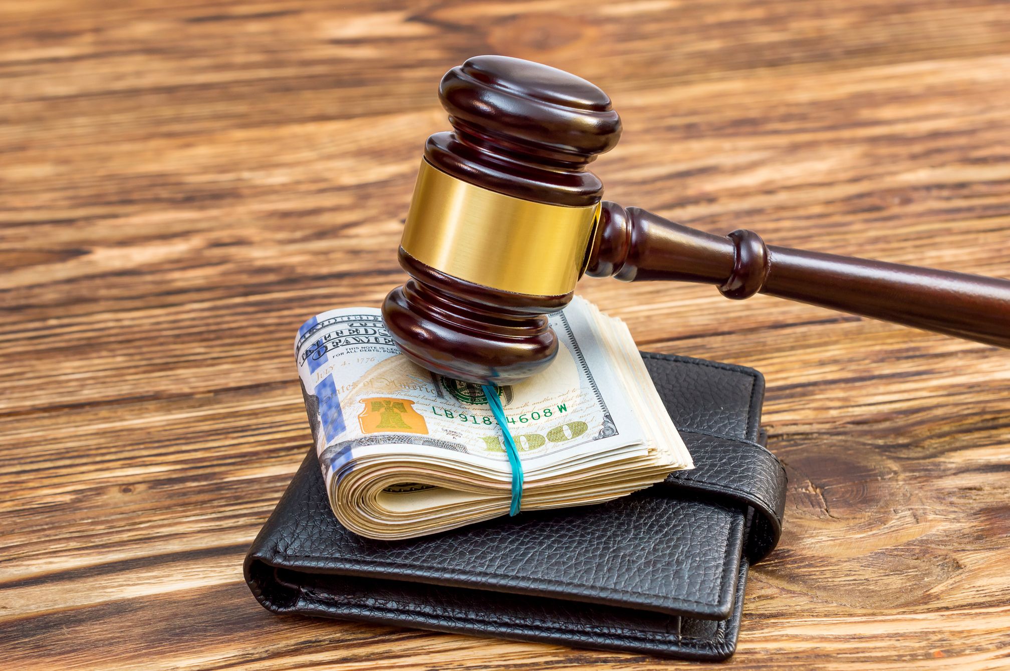 A wooden gavel rests on a bundle of U.S. dollar bills atop a black wallet.