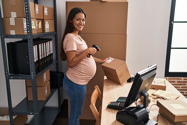 A pregnant worker uses a handheld scanner beside boxes and a computer in a stockroom.