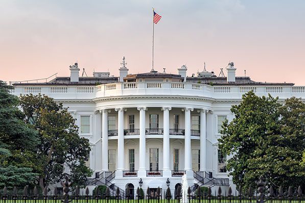 The White House beneath a pale pink sky with an American flag above it.