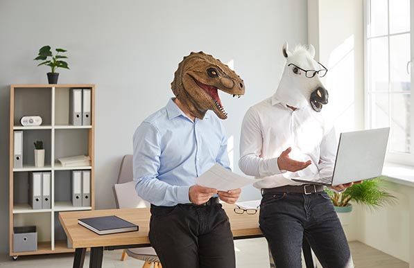 Two office workers wearing dinosaur and unicorn masks look at a laptop.
