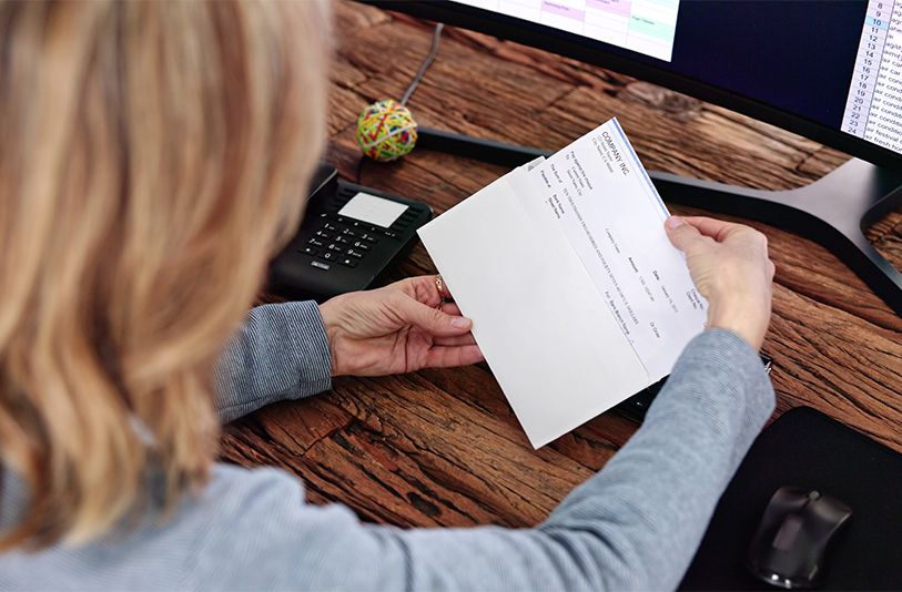 A person reviews a printed document at a desk beside a computer and telephone.