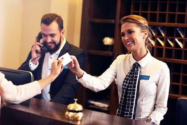 A smiling hotel receptionist hands a card to a guest while a colleague talks on the phone.