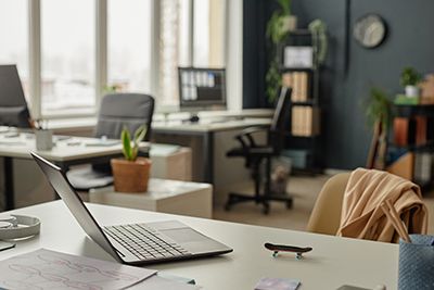 An open laptop sits on a desk in an empty, sunlit office.