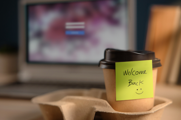 A takeaway coffee cup bears a yellow Welcome Back note with a smiley face in front of a laptop.