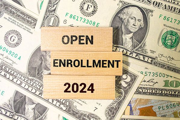 Wooden tiles reading Open Enrollment 2024 sit on overlapping U.S. dollar bills.