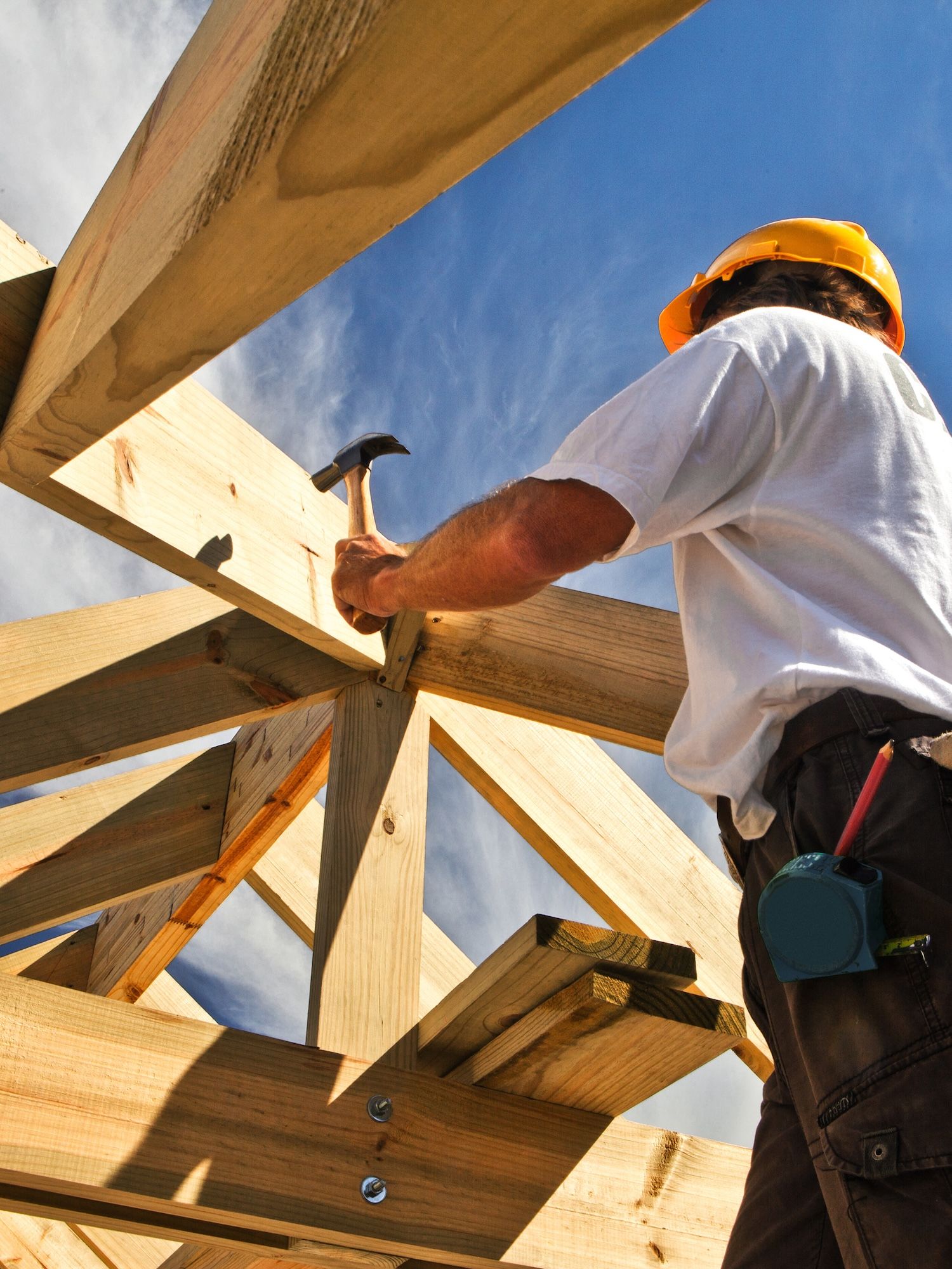 A construction worker in a hard hat uses a hammer on timber framing beneath a blue sky.