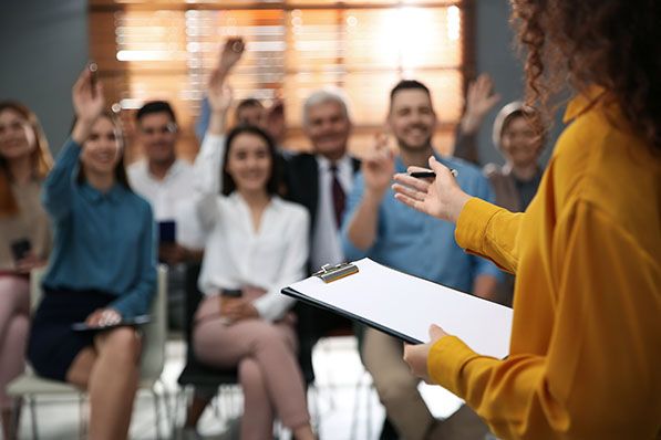 A presenter holding a clipboard addresses seated attendees, several with their hands raised.