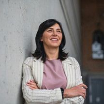 A smiling woman in a striped blazer stands with folded arms beside a concrete column.