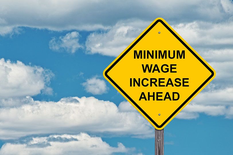 A yellow road sign reads Minimum Wage Increase Ahead against a cloudy sky.