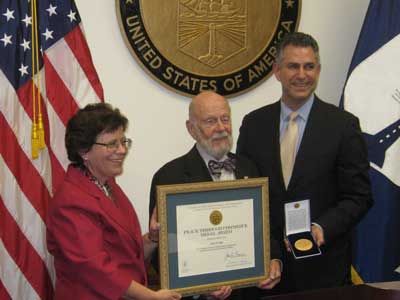 A woman and two men are shown with a certificate and medal