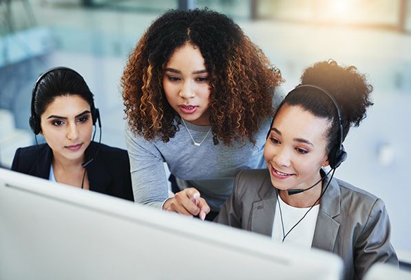 Three coworkers review a computer screen, two wearing headsets and one leaning in to assist.