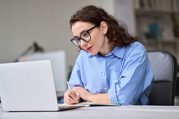 A woman wearing glasses writes in a notebook beside a laptop.