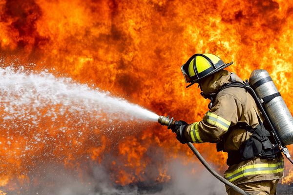 A firefighter in protective equipment sprays water toward a wall of flames.