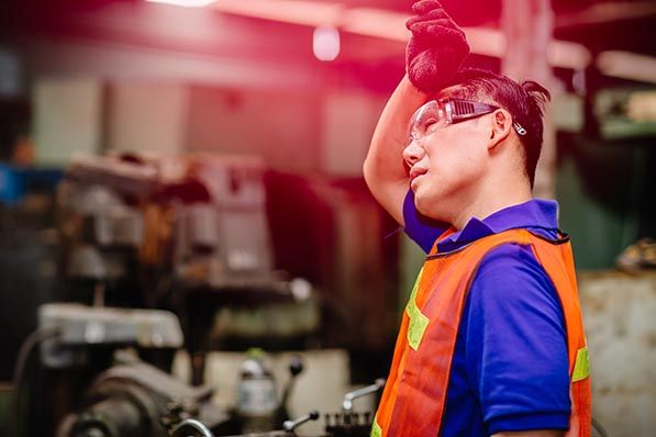 A worker in safety glasses and an orange vest raises a gloved hand to their forehead in a factory.