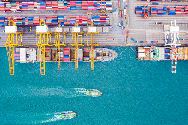 Overhead view of port cranes and rows of colorful shipping containers beside turquoise water and passing boats.