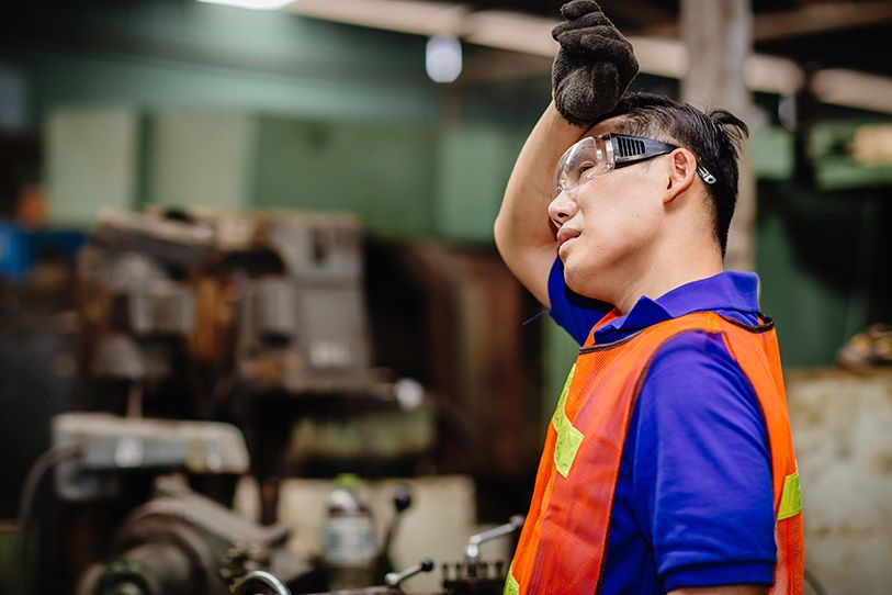 An industrial worker in an orange safety vest rests a gloved hand against their forehead.