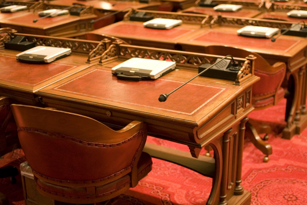 Empty wooden desks with microphones and leather chairs in a legislative chamber.