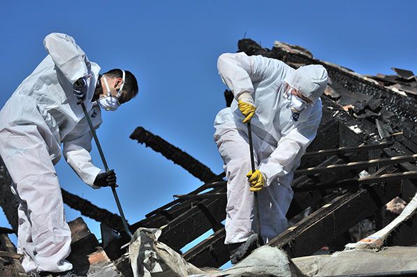 Two workers in white protective suits and masks use tools to clear charred debris.