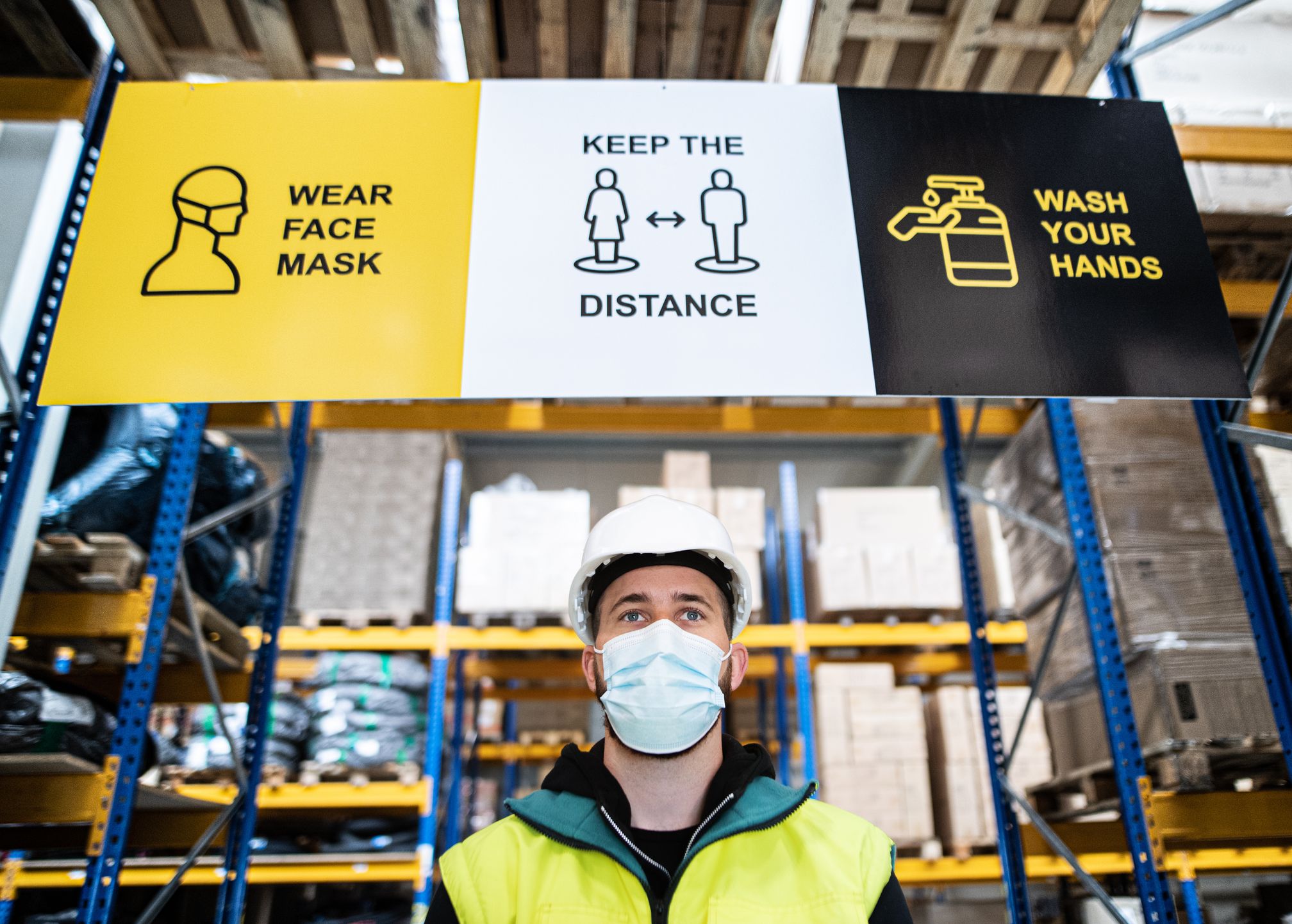 A masked warehouse worker stands beneath signs instructing people to wear a face mask, keep distance and wash hands.