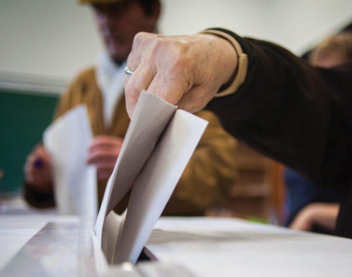 A hand lowers a folded ballot into a ballot box.
