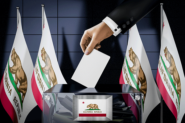 A hand lowers a ballot into a transparent box surrounded by California flags.